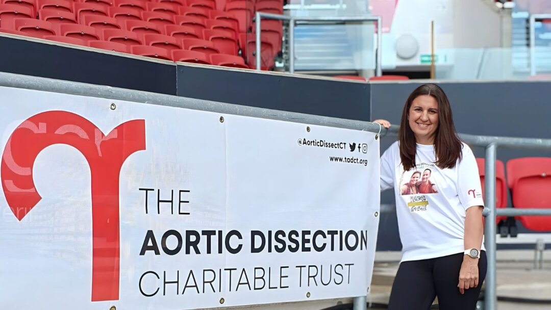 Charity supporter wearing a white T-shirt featuring a photo and text promoting aortic dissection awareness stands next to a large banner for The Aortic Dissection Charitable Trust. The woman is smiling, and the banner includes the charity's logo and social media handles, signifying participation in a fundraising or awareness campaign for aortic dissection.