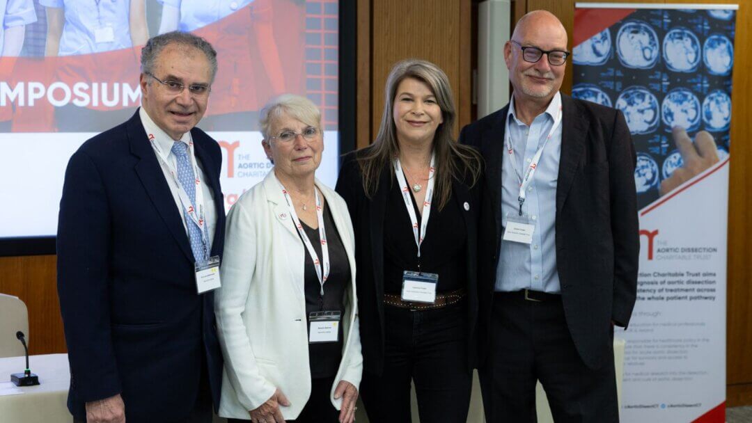 Four individuals, including Dr. John A. Elefteriades (on the left), stand together at an event for The Aortic Dissection Charitable Trust. They are wearing name badges and lanyards, smiling for the photo in front of a backdrop that references a symposium. A banner nearby outlines the charity's mission to improve diagnosis and treatment of aortic dissection. The setting is formal, and the group appears to be part of a significant moment marking a partnership related to the charity's work in aortic dissection awareness and patient care.