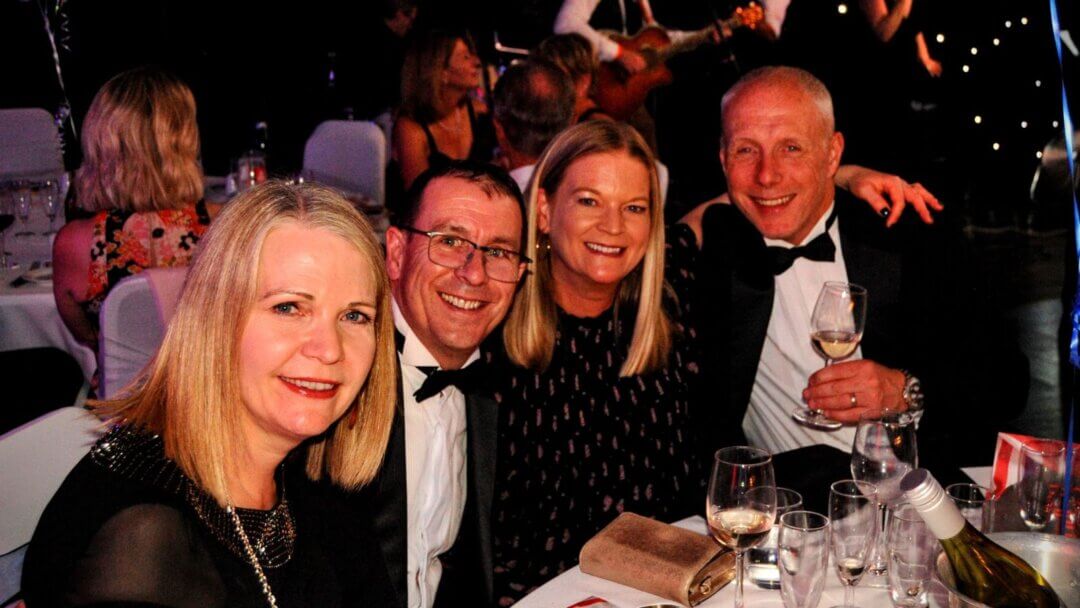 A lively moment at the Aortic Dissection Charity Ball, where four guests, dressed in formal attire, smile warmly at the camera. The elegant setting includes wine glasses and a bottle on the table, with a live band playing in the background, creating a festive and celebratory atmosphere in support of a meaningful cause.