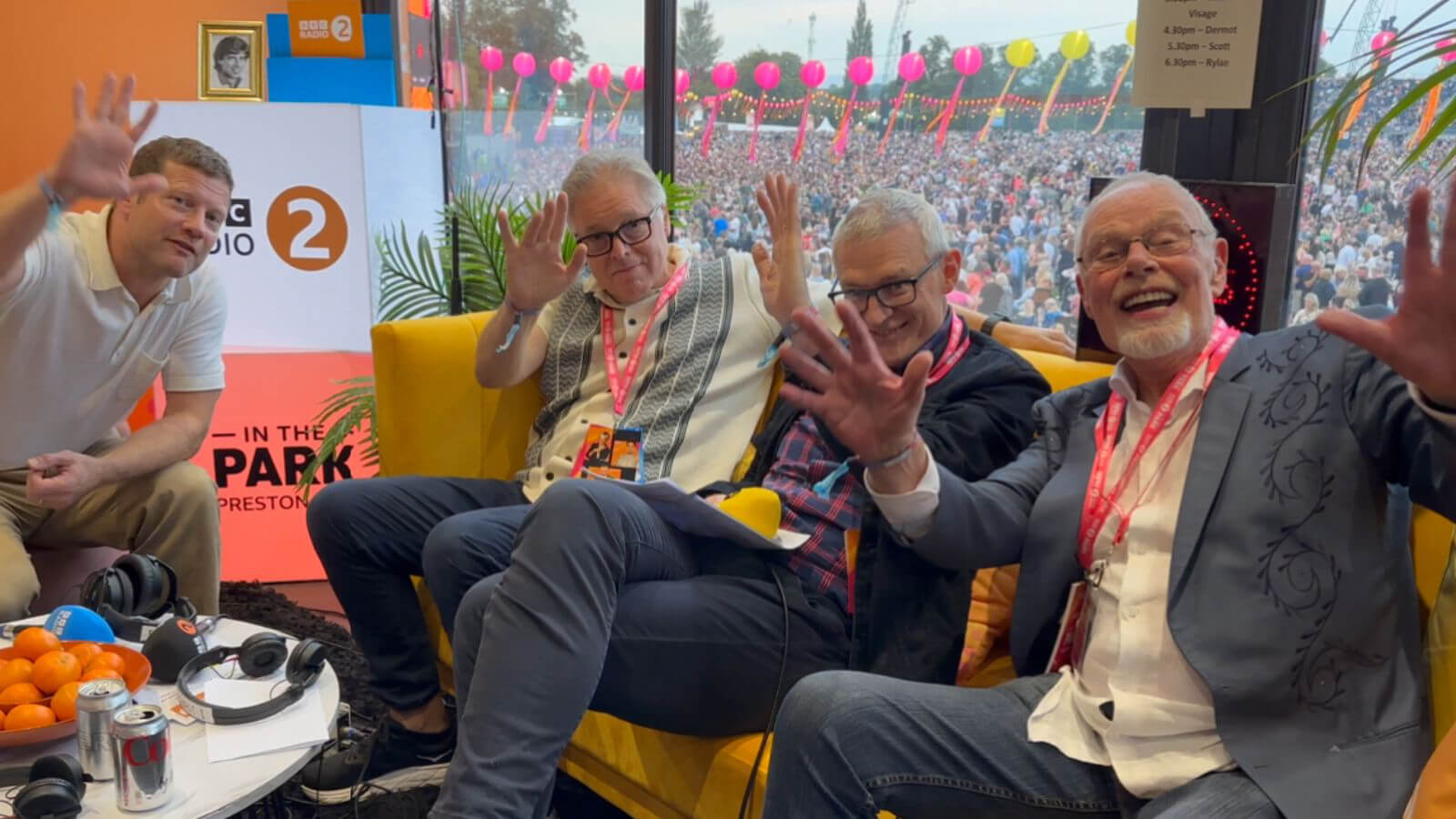 In this image, from left to right, Dermot O'Leary, Mark Goodier, Jeremy Vine, and Bob Harris are sitting on a yellow couch, smiling and waving towards the camera. Behind them, a BBC Radio 2 banner is displayed, and through the window, a large, festive crowd and vibrant pink balloons can be seen.